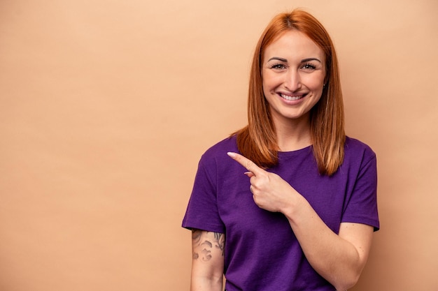 Young caucasian woman isolated on beige background smiling and pointing aside showing something at blank space