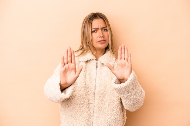 ベージュの背景に孤立した若い白人女性は、一時停止の標識を示している手を伸ばして立って、あなたを妨げています。