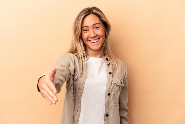 Foto giovane donna caucasica isolata su fondo beige che allunga la mano alla macchina fotografica nel gesto di saluto.