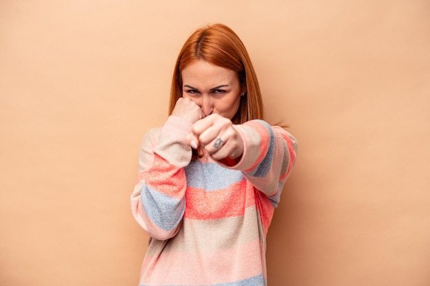 Young caucasian woman isolated on beige background throwing a punch, anger, fighting due to an argument, boxing.