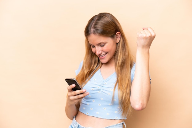 Young caucasian woman isolated on beige background with phone in victory position