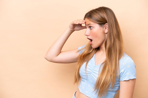 Young caucasian woman isolated on beige background with surprise expression while looking side