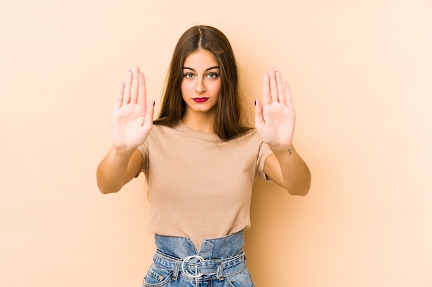 Young caucasian woman isolated on beige standing with outstretched hand showing stop sign, preventing you.