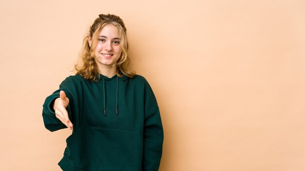 Young caucasian woman isolated on beige stretching hand in greeting gesture.