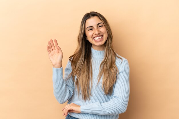 Young caucasian woman isolated on beige wall saluting with hand with happy expression