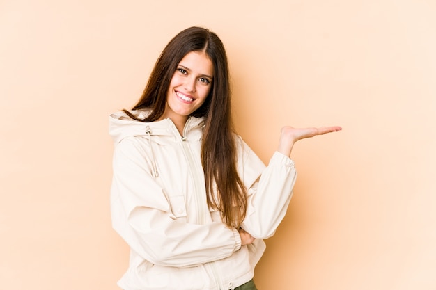 Young caucasian woman isolated on beige wall showing a copy space on a palm and holding another hand on waist.