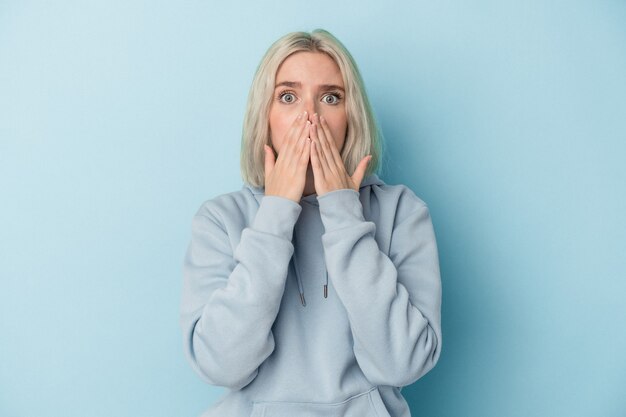 Young caucasian woman isolated on blue background covering mouth with hands looking worried.