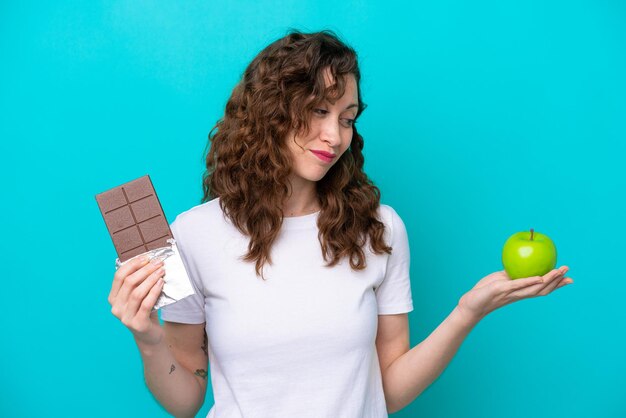 Young caucasian woman isolated on blue background having doubts while taking a chocolate tablet in one hand and an apple in the other