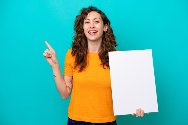 Young caucasian woman isolated on blue background holding an empty placard and pointing side