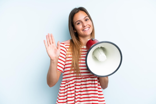 Photo young caucasian woman isolated on blue background holding a megaphone and saluting with hand with happy expression