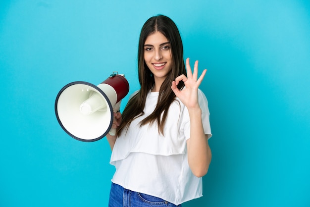 Young caucasian woman isolated on blue background holding a megaphone and showing ok sign with fingers