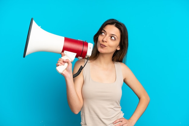 Young caucasian woman isolated on blue background holding a megaphone and thinking