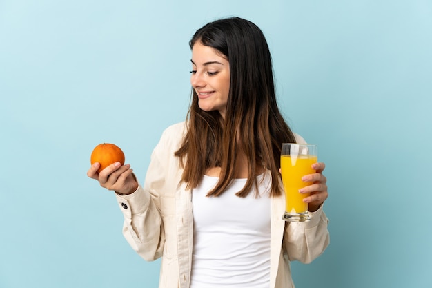 Молодая кавказская женщина изолирована на синем фоне, держа апельсин и апельсиновый сок