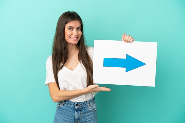 Young caucasian woman isolated on blue background holding a placard with arrow symbol with happy expression