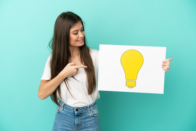 Young caucasian woman isolated on blue background holding a placard with bulb icon and pointing it