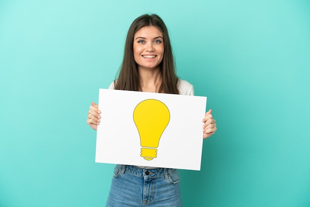 Young caucasian woman isolated on blue background holding a placard with bulb icon with happy expression