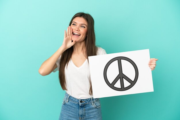 Young caucasian woman isolated on blue background holding a placard with peace symbol and shouting