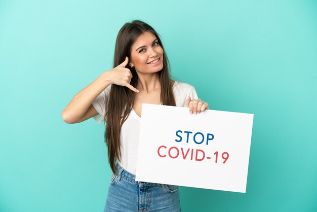 青の背景に分離された若い白人女性がテキストStopCovid 19のプラカードを保持し、電話ジェスチャーを行う