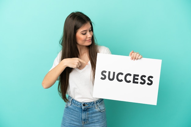 Young caucasian woman isolated on blue background holding a placard with text SUCCESS and  pointing it