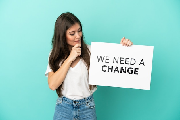 Young caucasian woman isolated on blue background holding a placard with text We Need a Change and thinking