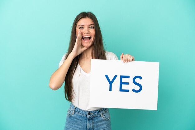 Young caucasian woman isolated on blue background holding a placard with text YES and shouting