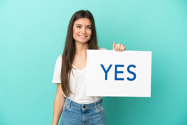 Young caucasian woman isolated on blue background holding a placard with text YES with happy expression