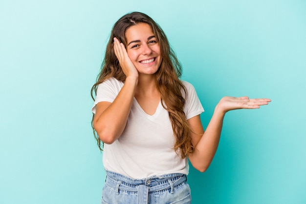 Young caucasian woman isolated on blue background  holds copy space on a palm, keep hand over cheek. Amazed and delighted.