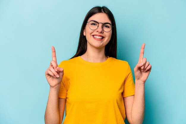 Young caucasian woman isolated on blue background indicates with both fore fingers up showing a blank space