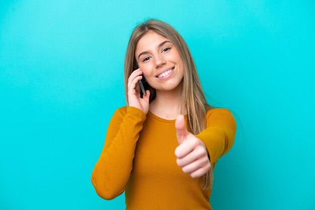 Young caucasian woman isolated on blue background keeping a conversation with the mobile while doing thumbs up