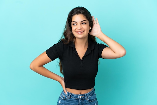 Young caucasian woman isolated on blue background listening to something by putting hand on the ear