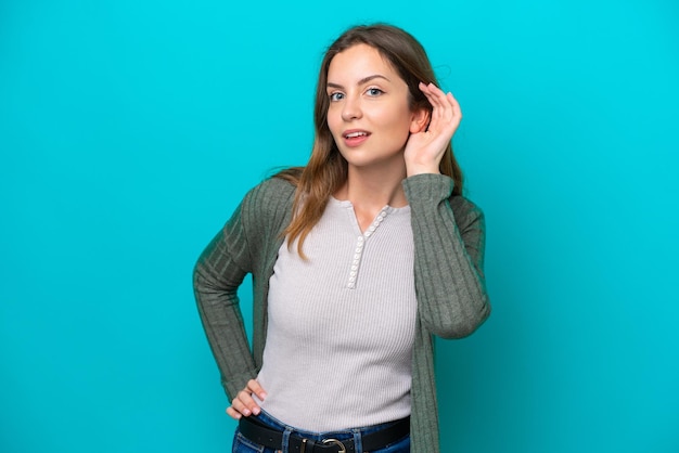 Young caucasian woman isolated on blue background listening to something by putting hand on the ear