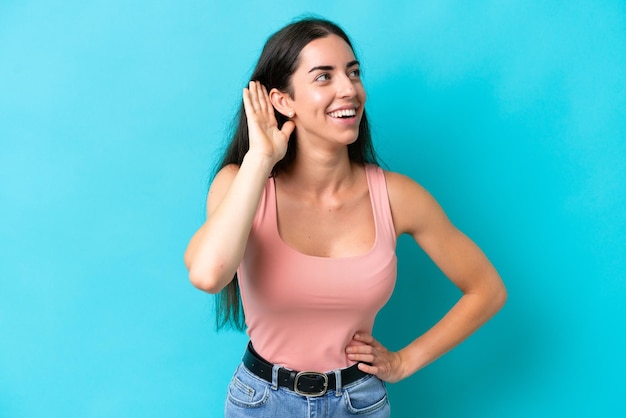 Young caucasian woman isolated on blue background listening to something by putting hand on the ear