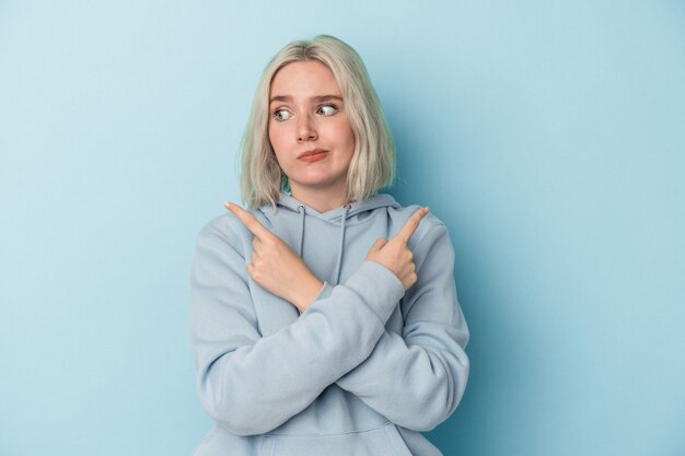 Young caucasian woman isolated on blue background points sideways, is trying to choose between two options.