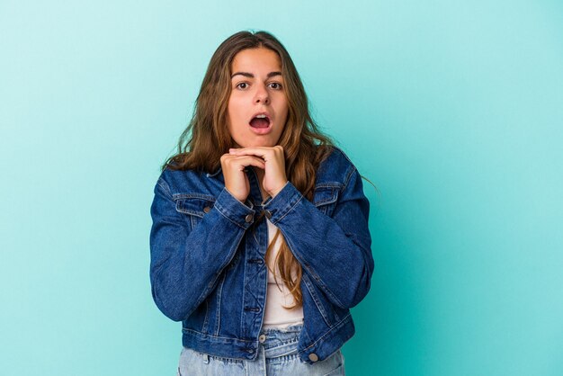 Young caucasian woman isolated on blue background  praying for luck, amazed and opening mouth looking to front.