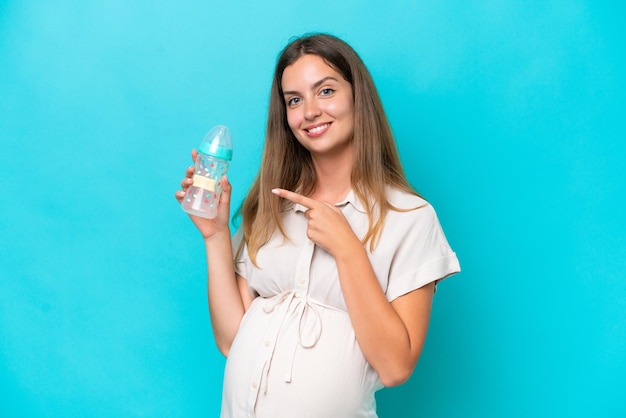 Young caucasian woman isolated on blue background pregnant and holding a feeding bottle