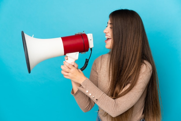 Young caucasian woman isolated on blue background shouting through a megaphone to announce something in lateral position