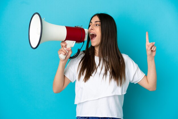 Young caucasian woman isolated on blue background shouting through a megaphone to announce something in lateral position