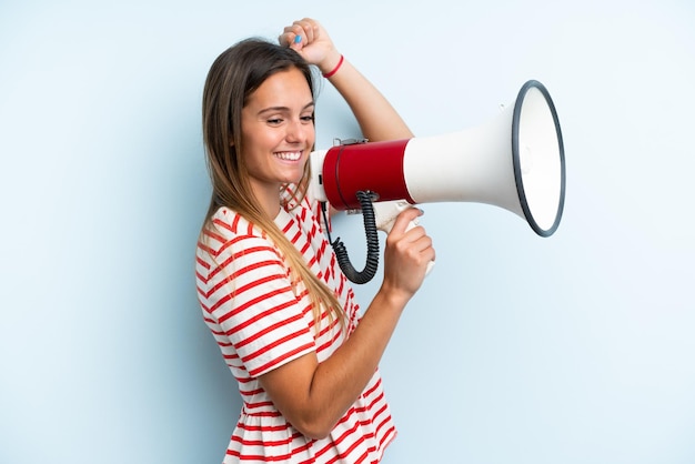 Young caucasian woman isolated on blue background shouting through a megaphone to announce something in lateral position