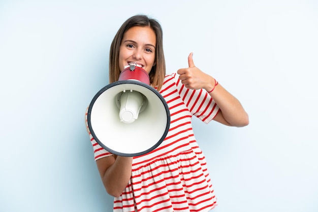 Young caucasian woman isolated on blue background shouting through a megaphone to announce something and with thumb up