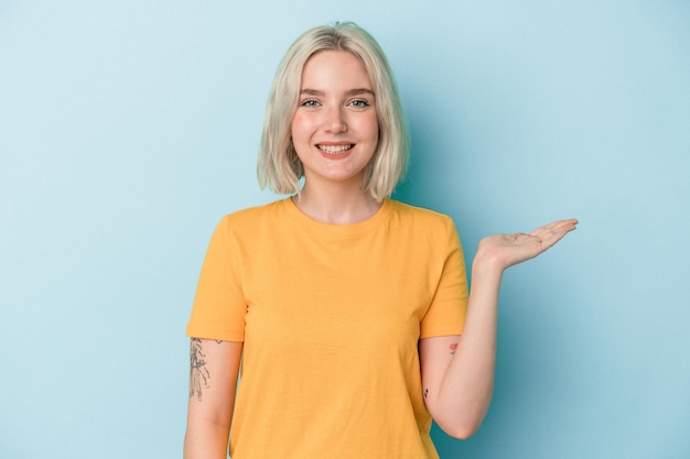 Young caucasian woman isolated on blue background showing a copy space on a palm and holding another hand on waist.