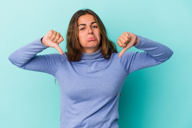 嫌いなジェスチャーを示す青い背景に分離された若い白人女性は、親指を下に向けます。不一致の概念。