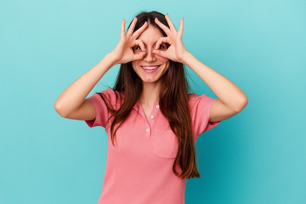 Young caucasian woman isolated on blue background showing okay sign over eyes