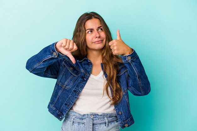 親指を上と親指を下に示す青い背景で隔離の若い白人女性、難しい選択の概念