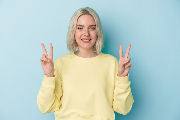 Young caucasian woman isolated on blue background showing victory sign and smiling broadly.