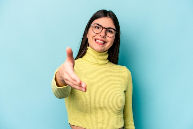 Foto giovane donna caucasica isolata su sfondo blu che allunga la mano alla telecamera nel gesto di saluto