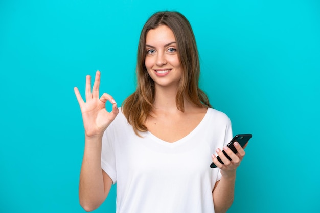 Young caucasian woman isolated on blue background using mobile phone and doing OK sign