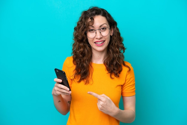Photo young caucasian woman isolated on blue background using mobile phone and pointing it