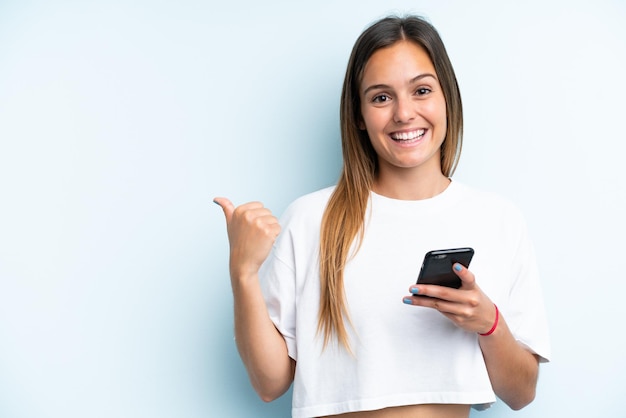 Young caucasian woman isolated on blue background using mobile phone and pointing to the lateral