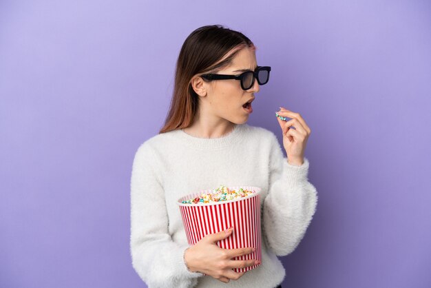 Young caucasian woman isolated on blue background with 3d glasses and holding a big bucket of popcorns while looking side