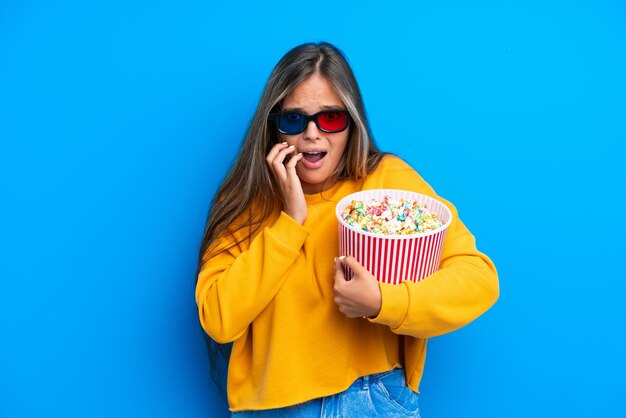 Young caucasian woman isolated on blue background with 3d glasses and holding a big bucket of popcorns